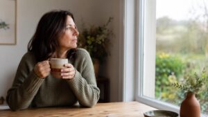 A woman sitting by a window with a cup of tea, pausing to reflect.